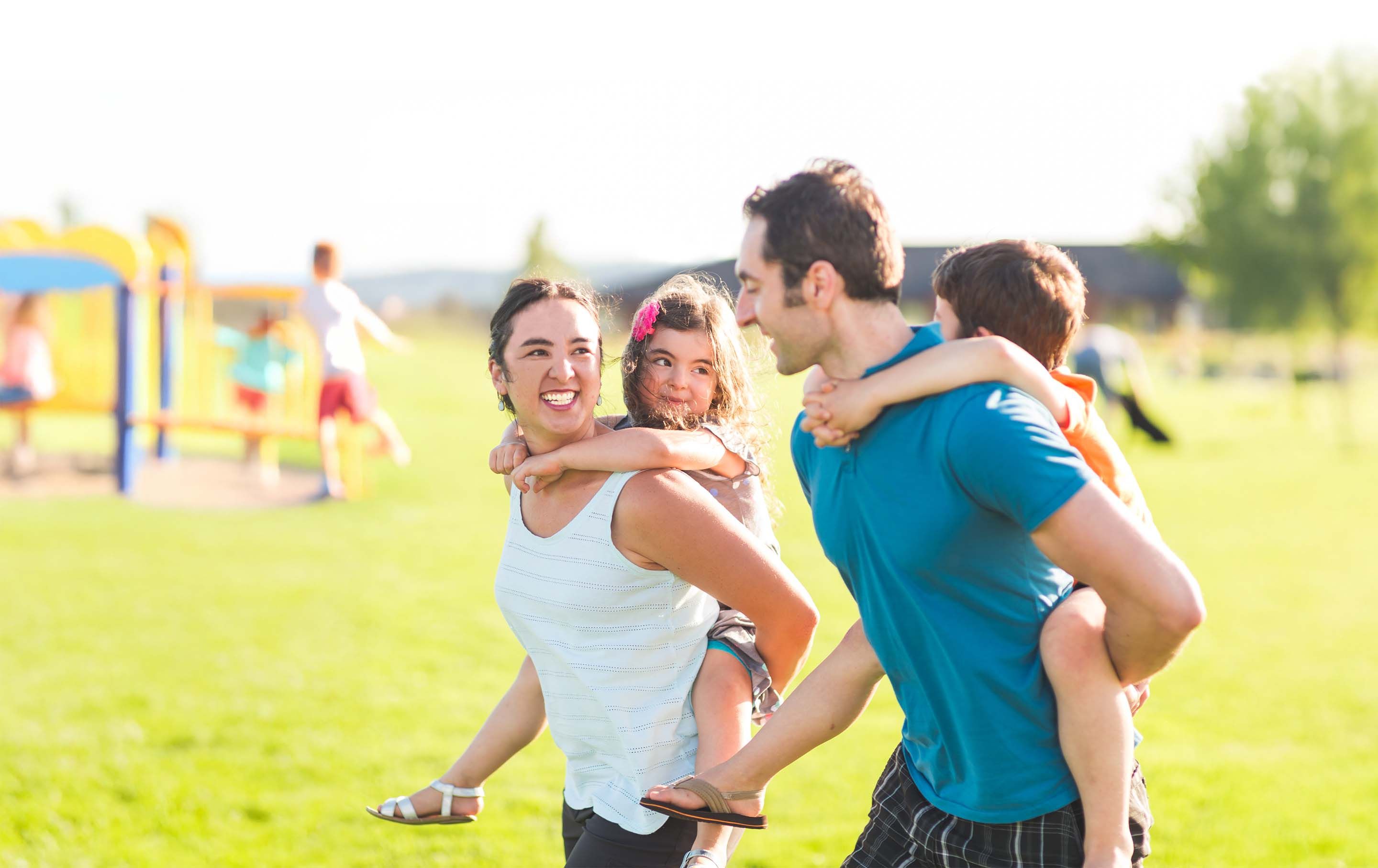 A cheerful mother and father give piggy-back rides to their young son and daughter while walking across a grassy park.