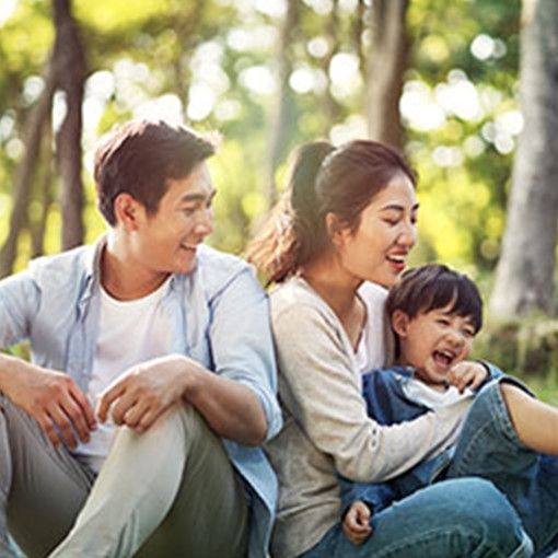 A family seated on the ground in a park, both parents look in the direction of a giggling child in the lap of the parent on the right.