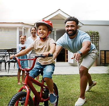 A young boy riding his bike in the backyard while being encouraged by his family.